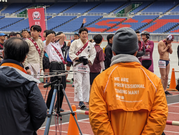 Eine Gruppe von Menschen, die auf einer Bahn stehen, mit einer Kamera auf einem Stativ im Vordergrund, Stühle, ein Banner und Lichter im Hintergrund.