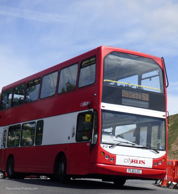 Ein roter Doppeldecker-Stadtbus fährt mit einem Verkehrskegel vorne und einem Hügel im Hintergrund.