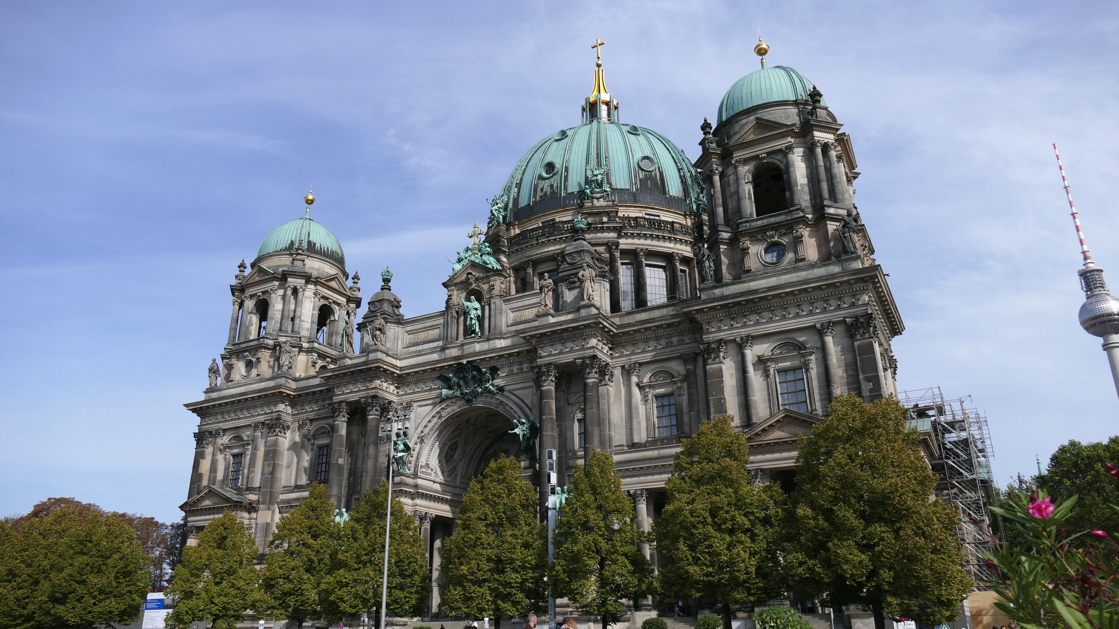 Der Berliner Dom, eine große Kathedrale mit prächtigen architektonischen Merkmalen, steht prominent in Berlin, Deutschland, mit Menschen in der Nähe und Bäumen, Blumenpflanzen und einem Turm im Vordergrund unter einem bewölkten Himmel.