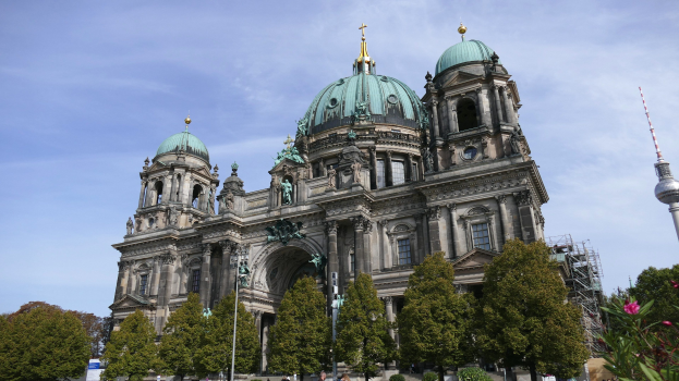 Der Berliner Dom, eine große Kathedrale mit prächtigen architektonischen Merkmalen, steht prominent in Berlin, Deutschland, mit Menschen in der Nähe und Bäumen, Blumenpflanzen und einem Turm im Vordergrund unter einem bewölkten Himmel.
