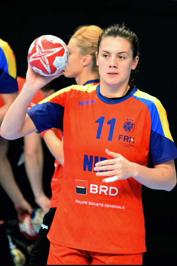 Eine Frau in einer orange-blauen Uniform hält einen Handball, schaut direkt in die Kamera und hat einen entschlossenen Gesichtsausdruck, ihre Haare sind zu einem Pferdeschwanz gebunden, mit ein paar Leuten im Hintergrund.