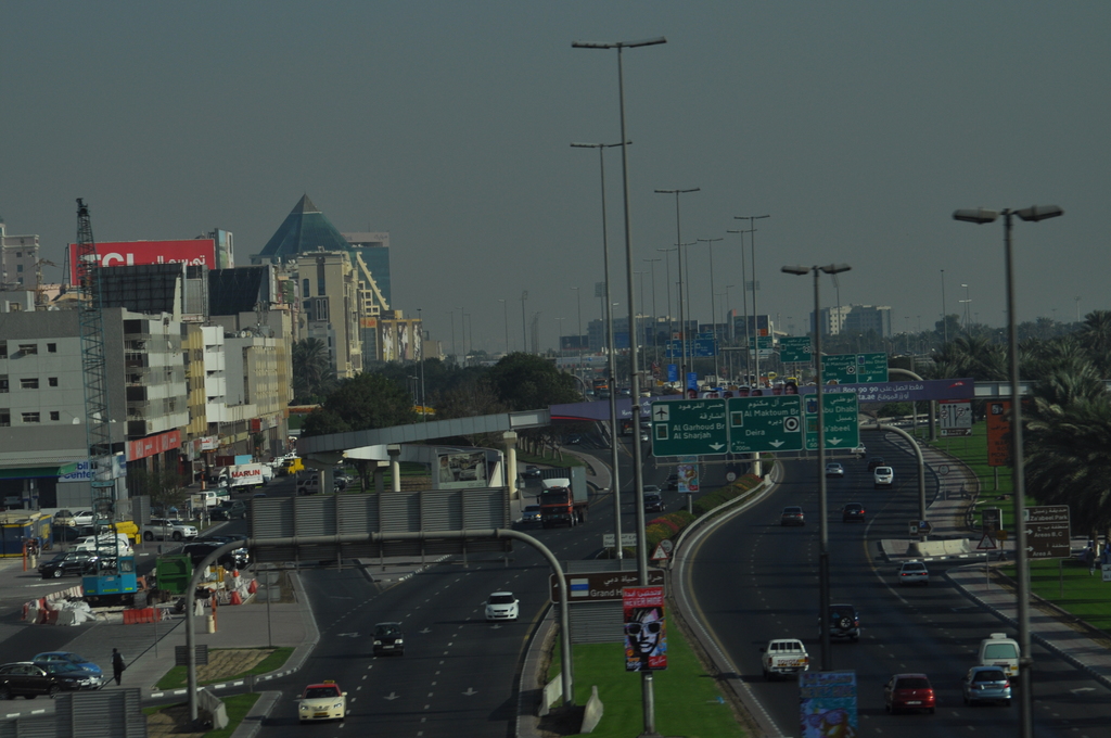 Eine Luftaufnahme einer Stadt mit Gebäuden, Türmen, Fahrzeugen, Werbetafeln, Schildern, Straßen, Bäumen, Straßenlaternen, Menschen, Verkehrsleitkegeln und Informationsschildern unter einem klaren Himmel.