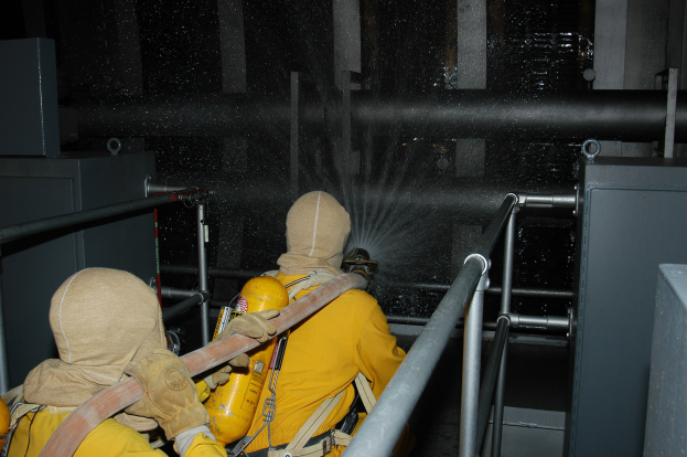 Zwei Feuerwehrleute in gelben Uniformen und Helmen spritzen Wasser aus einem Schlauch auf ein Metallgeländer, mit Metallstangen und anderen Gegenständen im Hintergrund und Text am unteren Bildrand.