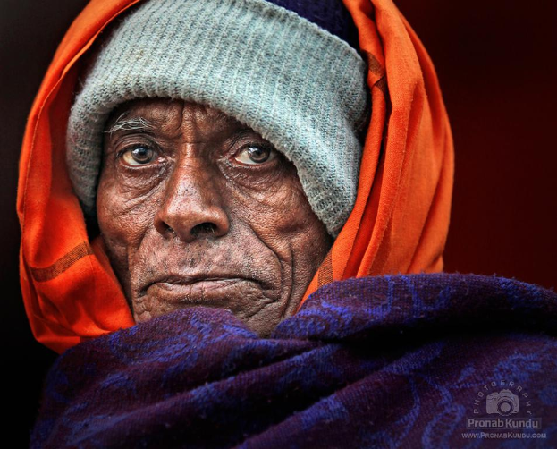 Ein älterer Mensch mit einer Mütze und einem orangen Tuch auf dem Kopf, der auf einem violetten Tuch steht, mit einem Logo in der unteren rechten Ecke.