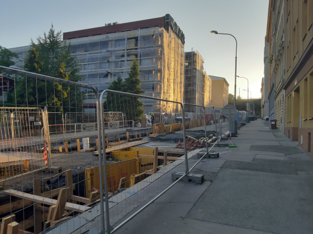 Eine Stadtstraße mit einer Baustelle in der Mitte, umgeben von Gebäuden, Straßenlaternen, Straßenlaternen, Fahrzeugen, Absperrpollern, Bäumen und einem klaren blauen Himmel.
