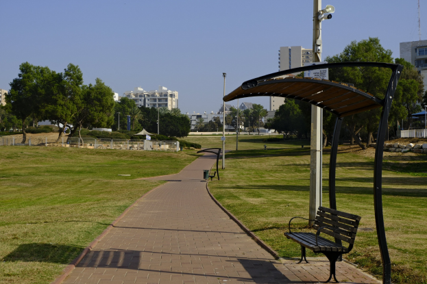 Eine Parkbank auf einer grünen Wiese umgeben von Bäumen, Mästen, Lichtern und Gebäuden, mit einem Mülleimer im Vordergrund.