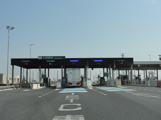 Mautstation mit einem vor ihr geparkten Lastwagen an einer Straße mit weißen und blauen Markierungen, Laternen, Schildern und Gebäuden, Bäumen und einem klaren blauen Himmel im Hintergrund.