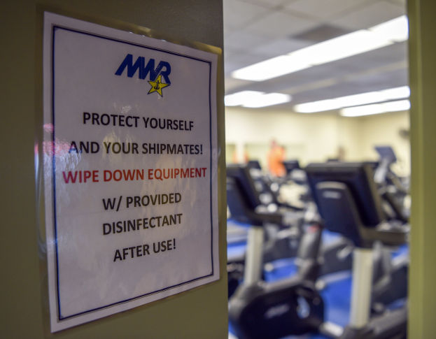 Ein Türschild mit der Aufschrift "Schützen Sie sich und Ihre Schiffskameraden" mit Laufbändern und Deckenleuchten im Hintergrund, das Bild leicht unscharf, um Bewegung zu suggerieren.