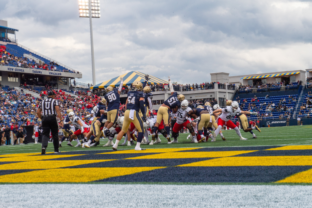 Ein Fussballspiel im Gange mit Spielern in Sportbekleidung und Helmen im Vordergrund, Zuschauern, Geländern, Beschriftungen, Laternenmasten und einem bewölkten Himmel im Hintergrund und Text am unteren Bildrand.