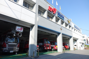 Eine Feuerwache mit mehreren Feuerwehrautos davor, mit Fenstern, Säulen und Namensschildern, Flaggen im Vordergrund und anderen Gebäuden unter einem klaren blauen Himmel im Hintergrund.