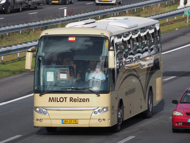 Ein Minibus mit Passagieren auf einer Autobahn neben einem roten Auto, mit Geländern und Gras am Straßenrand und Säulen mit Text im Hintergrund.