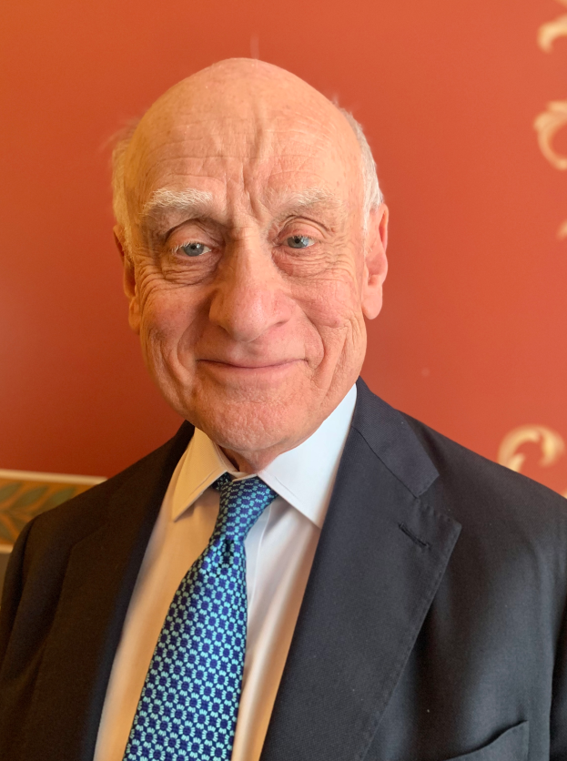 Ein älterer Mann im Anzug und Krawatte lächelt vor einer roten Wand, er scheint ein Nobelpreisträger zu sein.