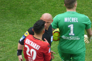 Schiedsrichter spricht mit zwei Fußballspielern auf einem saftig grünen Feld, ein Spieler hält einen Fußball.