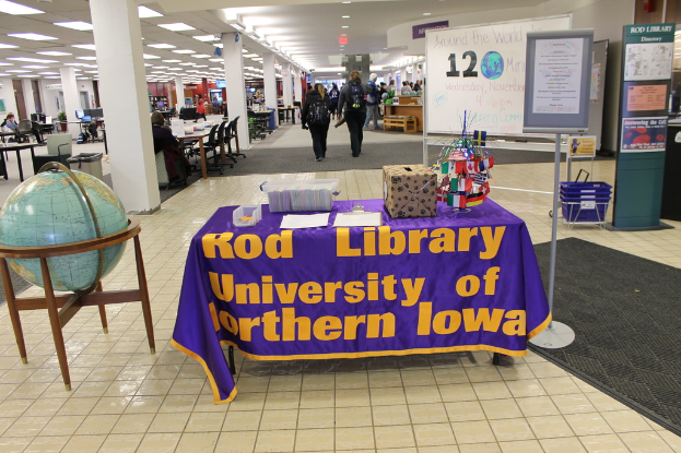 Eine Ausstellung mit einem Tisch, auf dem verschiedene Landesflaggen, eine Box, Papiere und ein Banner stehen, eine Kugel links daneben, ein Anschlagbrett rechts daneben mit ein paar Menschen in der Nähe und mehreren Tischen und Stühlen links.