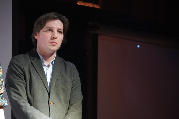 Zwei Personen nebeneinander auf einer Bühne, eine im Anzug, beide mit ernsten Gesichtern.