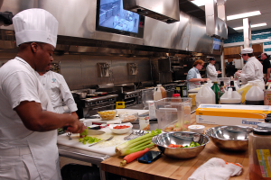 Eine Gruppe von Köchen in einer Küche beim Zubereiten von Essen auf einem Schneidbrett, wobei ein Koch im Vordergrund in einer weißen Schürze und einer Mütze steht und ein Messer hält und von verschiedenen Utensilien, Schüsseln, Gemüse, einem Handy, Flaschen und anderen Gegenständen umgeben ist, mit einem Fernseher und Deckenleuchten im Hintergrund.
