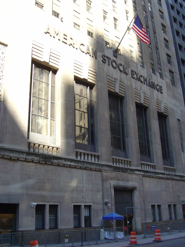 Ein Gebäude mit Text an seiner Fassade, ein Fahnenmast an der Wand, ein Geländer auf dem Gehweg davor, Absperrungen jenseits des Geländers und eine Straße im rechten unteren Bereich.