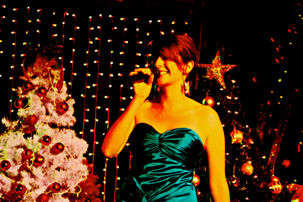 Eine Frau steht mit einem Mikrofon in der Hand vor Weihnachtsbäumen und Lichtern.