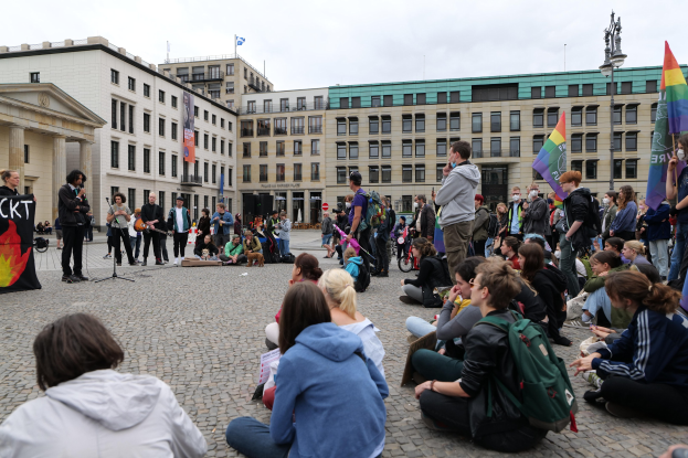 Eine Gruppe von Menschen, die auf dem Boden vor einer Menge sitzen, die Fahnen und Plakate hält, mit einer Person, die in ein Mikrofon spricht, einer Statue und Gebäuden im Hintergrund während einer Demonstration gegen Homosexuelle in Berlin.