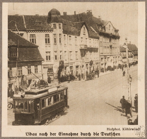 Ein altes Schwarz-Weiß-Foto einer Straße mit einer Tram, Gebäuden, Straßenmästen, Straßenlaternen und Passanten, mit Text unten.