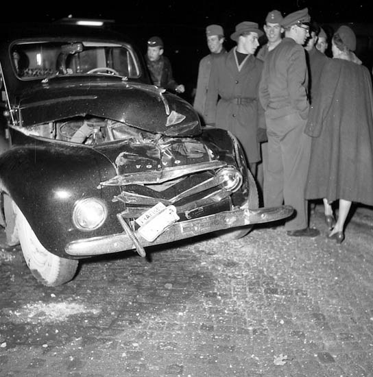 Schwarz-weißes Bild eines schwer besch"adigten Autos am Straßenrand mit einer Gruppe von Menschen, die es inspizieren.
