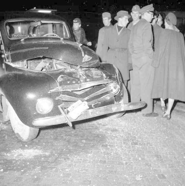 Schwarz-weißes Bild eines schwer besch"adigten Autos am Straßenrand mit einer Gruppe von Menschen, die es inspizieren.