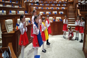 Eine Gruppe von Kindern hält Fahnen und steht vor einem großen Raum mit Stühlen, Tischen und anderen Gegenständen, mit einigen Menschen, die sitzen und stehen, und einer Tür und einer Treppe auf der rechten Seite.