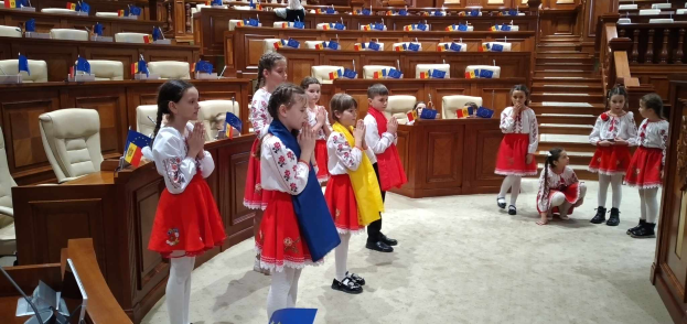 Eine Gruppe von Kindern hält Fahnen und steht vor einem großen Raum mit Stühlen, Tischen und anderen Gegenständen, mit einigen Menschen, die sitzen und stehen, und einer Tür und einer Treppe auf der rechten Seite.