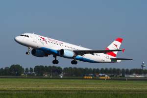 Austrian Airlines Airbus A320-200 starting from Frankfurt Airport, with green grass below, trees and buildings in the background, and a tower on the right.
