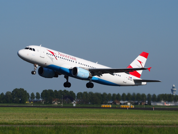 Austrian Airlines Airbus A320-200 starting from Frankfurt Airport, with green grass below, trees and buildings in the background, and a tower on the right.
