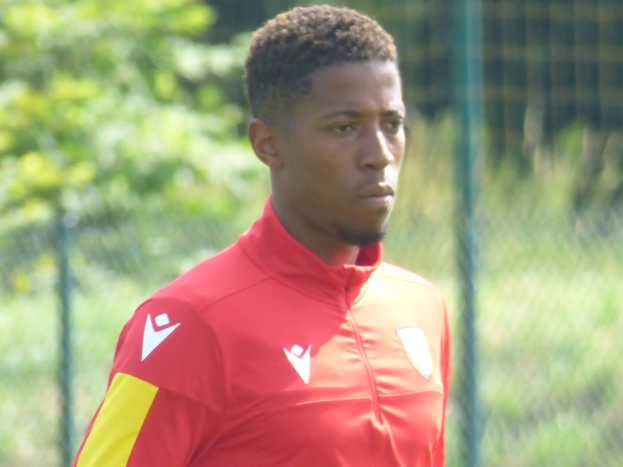 Ein junger Mann in einem roten und gelben T-Shirt steht auf einem Fußballfeld umgeben von einem Zaun und Bäumen und sieht entschlossen aus während eines Trainings.