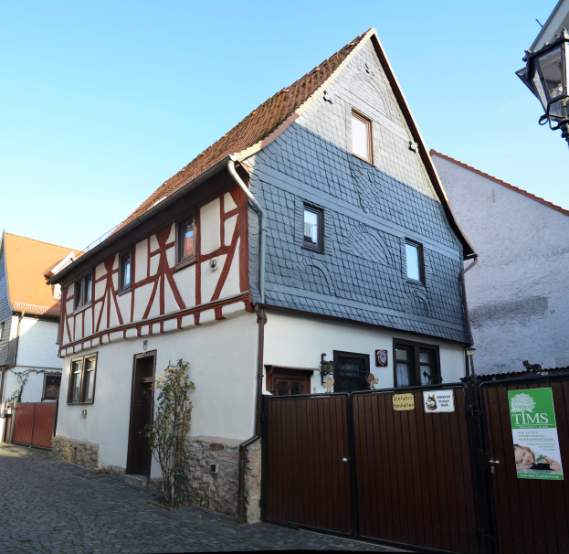 Ein Fachwerkhaus in Rothenburg ob der Tauber, Deutschland, mit Fenstern, Türen, einem hölzernen Tor mit Plakaten, einer Pflanze und einem Laternenmast, mit Text unten und dem Himmel im Hintergrund.