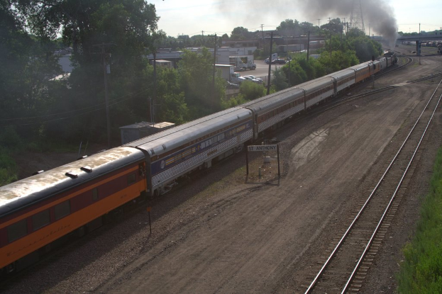 Ein Zug auf Schienen mit Bäumen, Strommasten mit Kabeln, Autos und Gebäuden links daneben und eine Brücke davor, aus der Rauch aufsteigt.