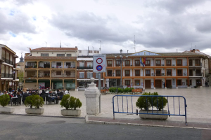 Ein belebter Stadtplatz mit Menschen auf Stühlen und Stehenden, umgeben von Topfpflanzen, Metallabsperrungen, Straßenmasten mit Flaggen, einem Schild, Gebäuden mit Fenstern und einem bewölkten Himmel.
