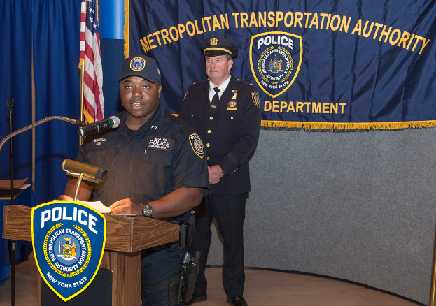 Zwei Polizeibeamte stehen vor einem Rednerpult, einer hält Papiere und spricht in ein Mikrofon, mit einer Fahne, einem Tisch und einer Tafel mit Symbolen und Text in der Nähe