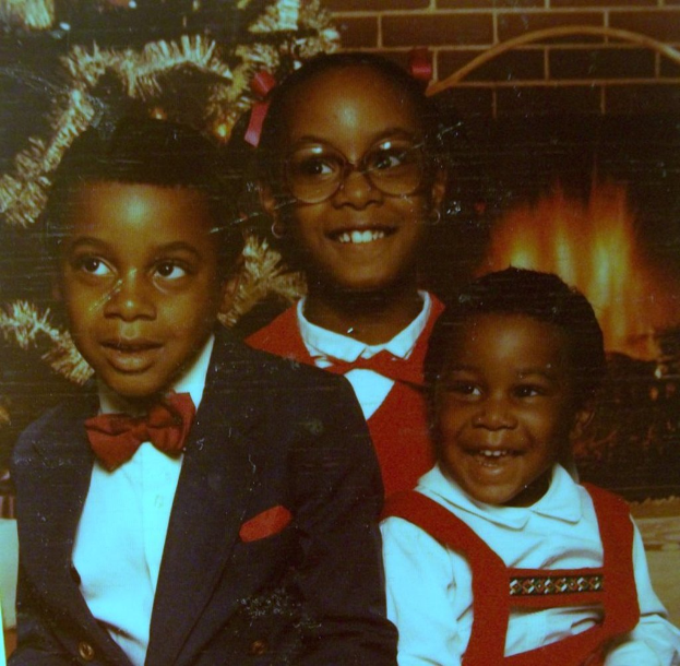 Drei Kinder, die vor einem Weihnachtsbaum mit einem Kamin aus Holzscheiten auf der rechten Seite lachen.