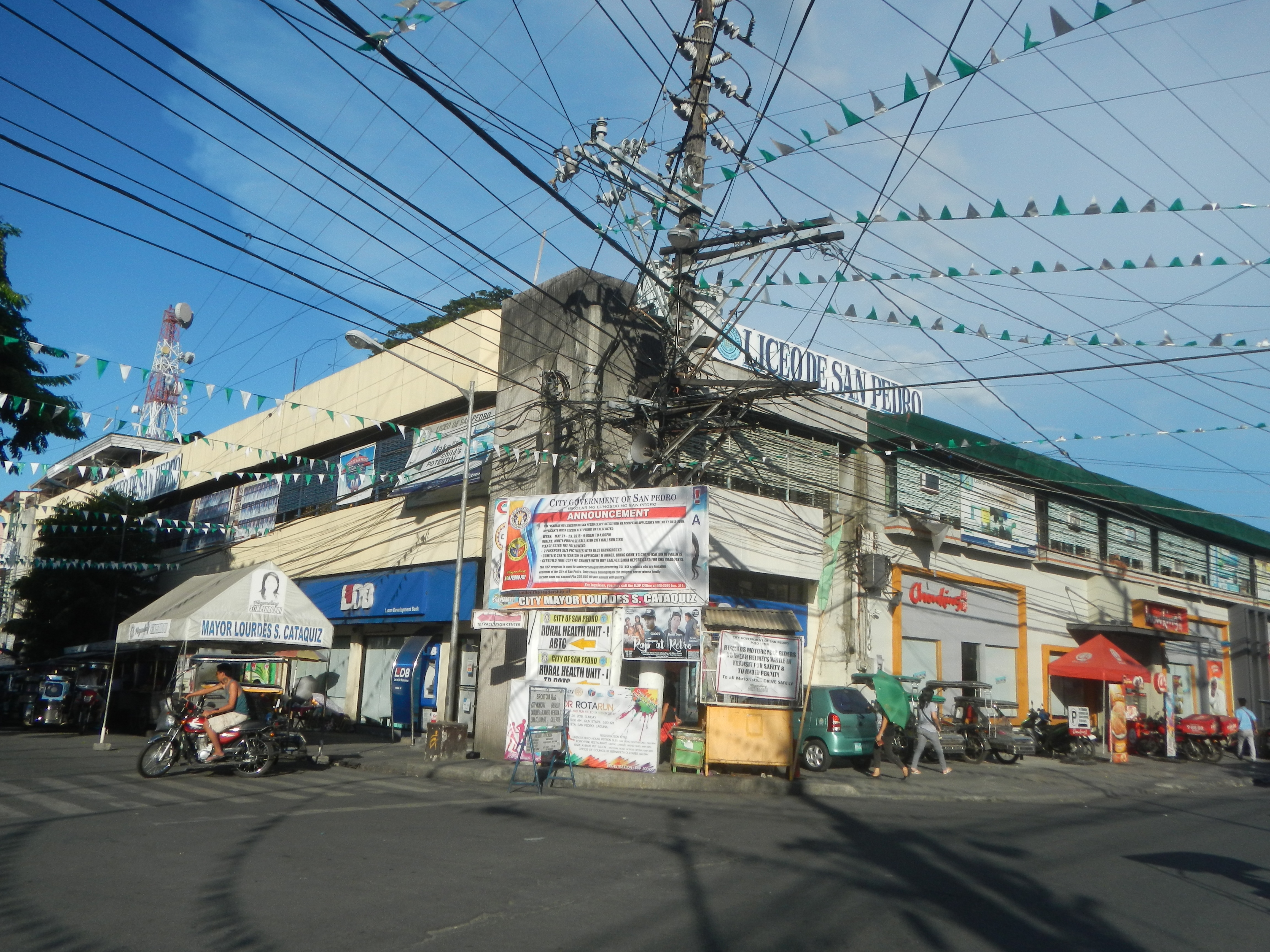 Eine Stadtstraßenszene mit einem Telefonmast in der Mitte, umgeben von Gebäuden, Geschäften, Fahrzeugen, Fußgängern, Schirmen, Strommasten, Kabeln, Fahnen, Bäumen und einem bewölkten Himmel.