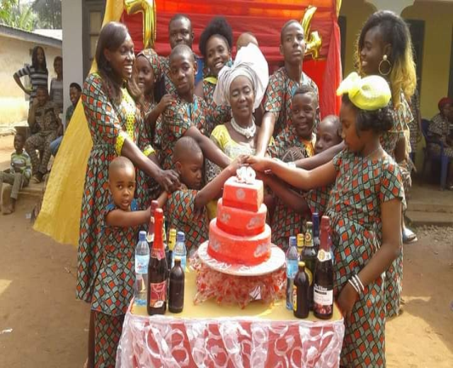 Eine Gruppe von Menschen sitzt um einen Tisch mit einem Kuchen, Flaschen und anderen Gegenständen in einer ländlichen Umgebung mit Häusern, Bäumen und einem klaren blauen Himmel, was auf eine Geburtstagsfeier hindeutet.