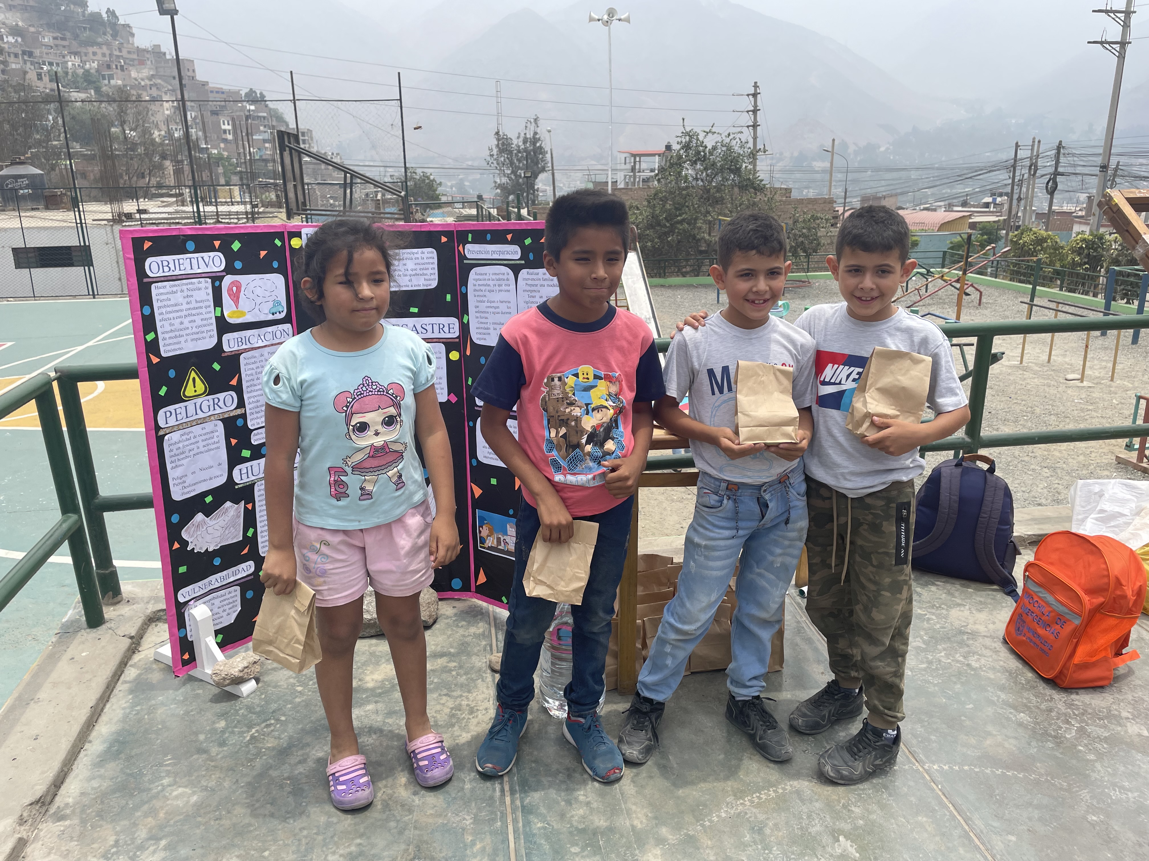 Eine Gruppe von Kindern steht auf einem Basketballplatz, jedes hält einen Papiertüte, mit Tüten und Gegenständen rechts und einer Tafel mit Text links, mit Zaun, Pfosten, Straßenlaternen, Bäumen, Gebäuden, Hügeln und Himmel im Hintergrund.