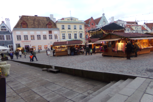 Ein lebendiger Weihnachtsmarkt in Tallinn, Estland, mit Menschen um geschmückte Stände, festlicher Beleuchtung und Gebäuden mit Fenstern im Hintergrund, unter einem bewölkten Himmel, mit Stufen, die zum Markt führen und Töpfen mit Pflanzen, die herumstehen.