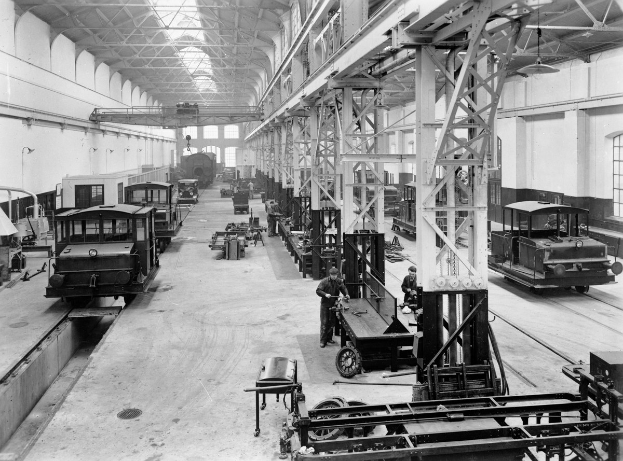 Schwarzes und weißes Foto einer Fabrikhalle mit mehrerenbahnen in Reparatur, Arbeitern, die sich darum kümmern, Metallpfosten, die das Dach stützen, Deckenlampen und Fenstern an den Wänden.