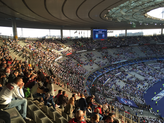 Eine große Menschenmenge sitzt im Allianz Arena Stadion in München, Deutschland, bei einem Fußballspiel, mit einer Bühne auf der rechten Seite, Fahnen, Stangen und einem Bildschirm im Hintergrund, unter einem sichtbaren Himmel.