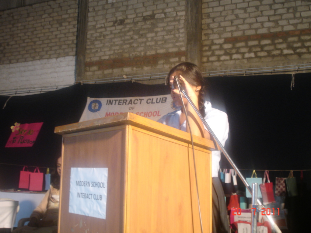 Ein Mädchen in einem weißen Hemd steht in der Nähe eines Rednerpults in einem Konferenzraum, mit einem Mikrofonständer zu seiner Rechten, einem weißen Banner auf einem Hintergrund hinter ihm und mehreren Taschen, die an einem Faden zu seiner Rechten hängen.