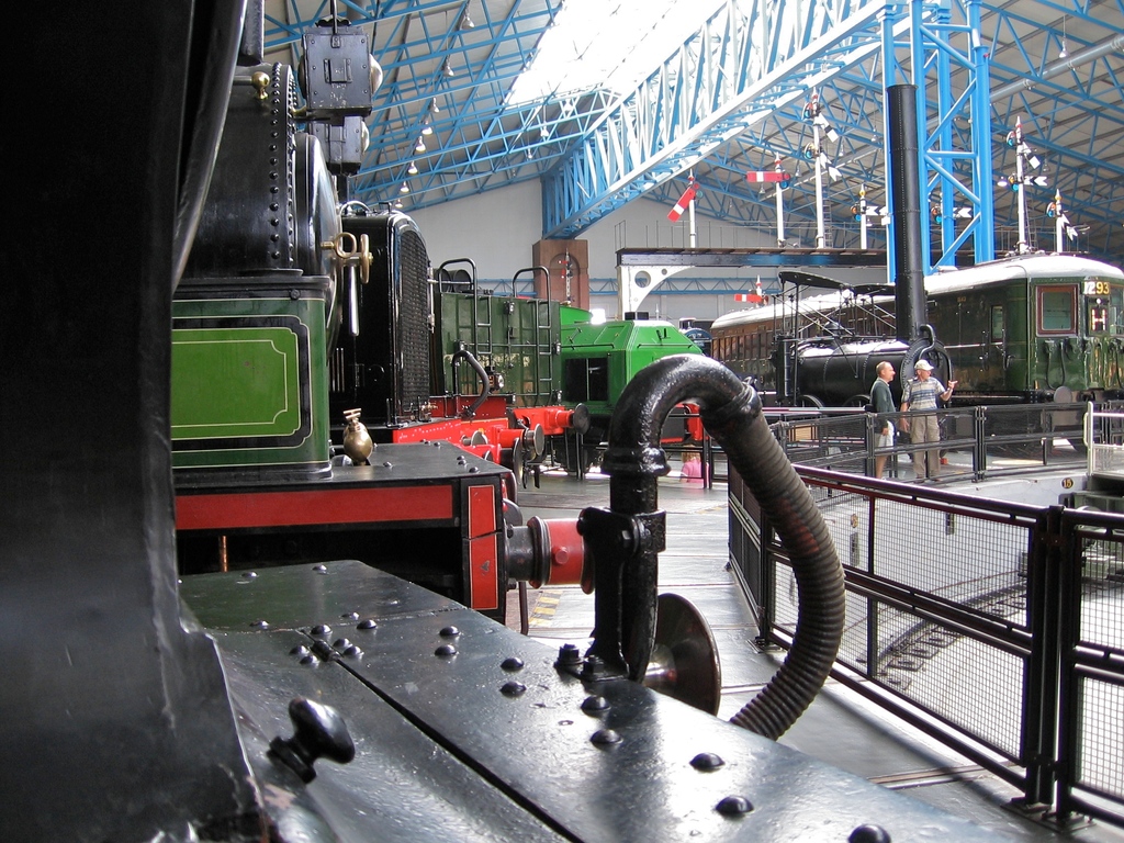 Mehrere Zugmaschinen stehen in einem Abstellgleis mit zahlreichen Eisenstangen darüber, einem Zaun auf der rechten Seite, einem Zug auf dem Bahngeleis neben dem Zaun und zwei Personen in der Mitte nahe dem Zaun.