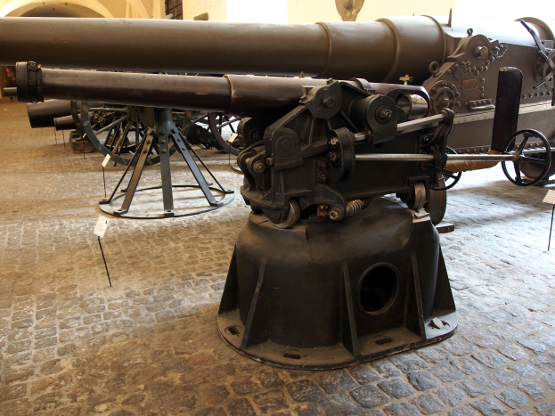 Eine große Gatling-Kanone mit langem Lauf und einem Seitengriff ist in einem Museum ausgestellt, umgeben von Bodenständen, mit einer fensterumgebenen Wand im Hintergrund, die an eine Burg erinnert.