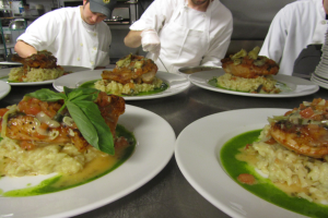 Drei Köche in einer Küche mit Mützen und Handschuhen bereiten Teller mit Essen zu, mit Gestellen im Hintergrund.