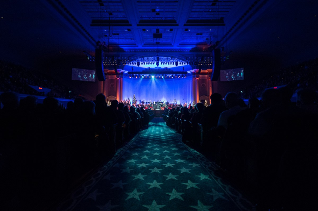 Ein großer Saal mit sitzenden Zuschauern, eine Bühne mit Musikern und eine Decke mit Lichtern und Fernsehgeräten, was auf eine Konzerthalle für die Nationale Convention 2019 hinweist.