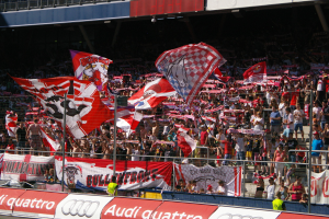 Eine große Menschenmenge in einem Stadion mit Fahnen, Bannern, einem Metallzaun, einem Schild, einem Anzeigebildschirm und Deckenleuchten.