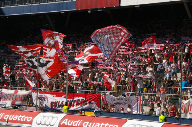 Eine große Menschenmenge in einem Stadion mit Fahnen, Bannern, einem Metallzaun, einem Schild, einem Anzeigebildschirm und Deckenleuchten.