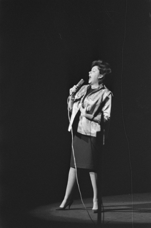 Eine Frau in einem schwarz-weißen Outfit aus den 1950er-Jahren singt fröhlich auf einer von einem Scheinwerfer beleuchteten Bühne und hält ein Mikrofon in der Hand.